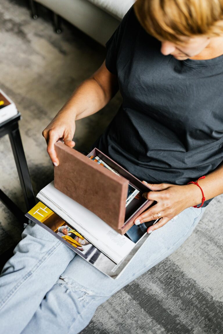 A woman in denim jeans sits indoors organizing a photo box beside magazines.